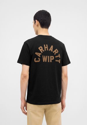 Uomo che indossa una maglietta nera a maniche corte con testo marrone "CARHARTT WIP" sulla schiena, abbinata a pantaloni beige, in piedi di spalle.