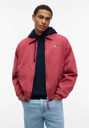 Ung mand iført en rød lynlåsjakke over en mørkeblå hættetrøje og lyseblå jeans, stående med hænderne i jakkelommerne mod en hvid baggrund.