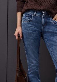 Jeans de mezclilla azul con un acabado ligeramente desgastado, diseño de cintura alta, con dos bolsillos delanteros, cierre de botón y un bolso de ante marrón.