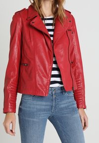 Giacca da biker in pelle rossa con un design aderente, zip asimmetrica e dettagli a coste sulle maniche, abbinata a una maglietta a righe e jeans blu.