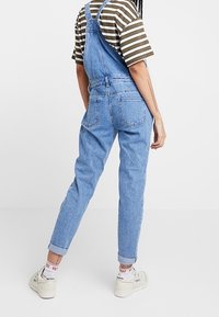 Salopette en denim bleu clair avec des bretelles réglables, des jambes fuselées et des ourlets roulés, associée à un t-shirt rayé brun et crème.