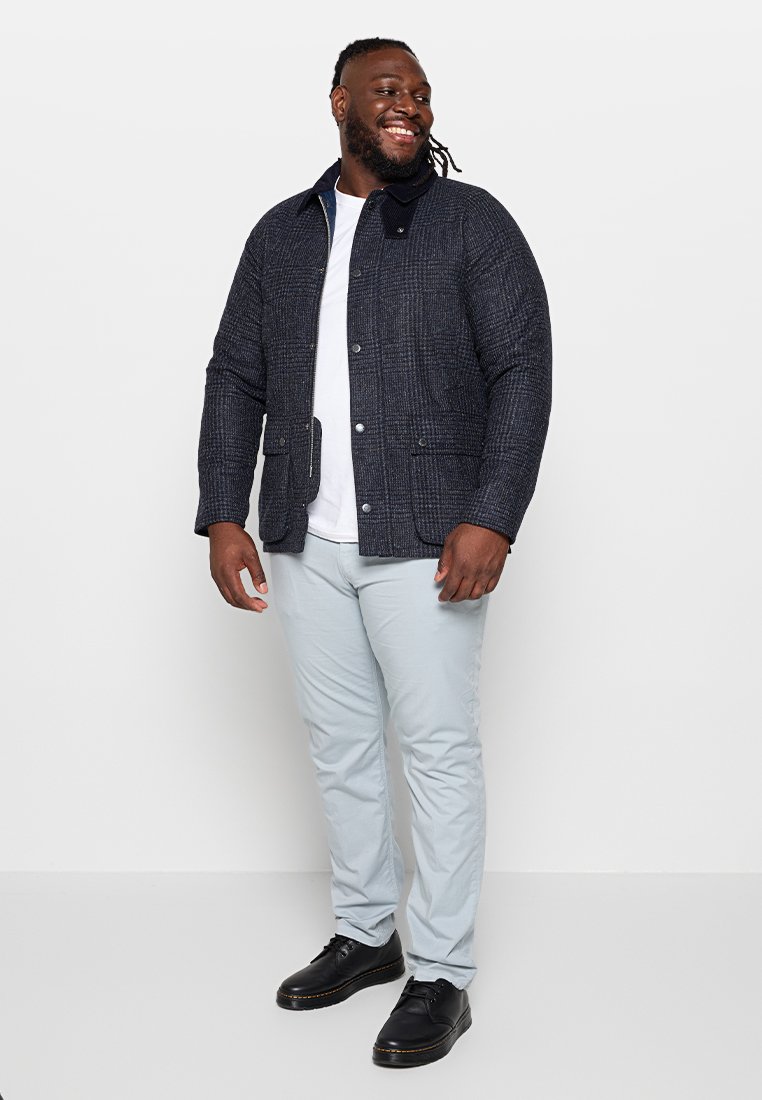 Mann mit Dreadlocks, der lächelt, trägt eine dunkelblaue gemusterte Jacke, ein weißes T-Shirt, hellgraue Hosen und schwarze Schuhe, steht vor einem weißen Hintergrund.