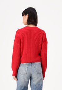 Rode gebreide trui met een losse pasvorm, verlaagde schouders en geribde cuffs, gecombineerd met lichtblauwe jeans met stervormige versieringen.