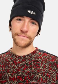 Beanie di pile nero con logo circolare; shirt di pile con motivo rosso, nero e beige, con colletto rotondo e tessuto testurizzato.