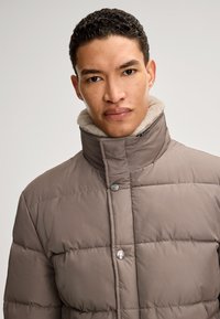 Pufferjack met hoge kraag in taupe, voorzien van een zachte fleece voering en metalen drukknoopsluitingen. Ontworpen met een geborduurd patroon.