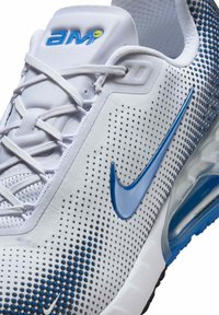 White athletic shoe with blue accents, perforated texture, and a distinct Nike swoosh. Features a cushioned sole and lace-up design.