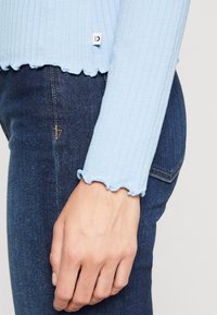 Himmelblaues, geripptes Langarmoberteil mit gewelltem Saum und Bündchen, kombiniert mit dunklen Denim-Jeans. Das Outfit zeigt einen strukturierten Stoff und eine lässige Passform.