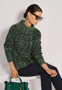 Frau in einem schwarzen, grünen und blauen Strickpullover, schwarzen Hosen, großen goldenen Creolen, schwarzen Sonnenbrillen, die eine grüne Handtasche hält und sitzt.