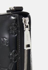 Pochette in pelle verniciata nera con zip, decorata con modelli floreali in rilievo, hardware argentato e una texture liscia. Presenta un evidente dettaglio della chiusura a zip.