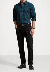 Grün- und blau kariertes Button-Up-Hemd, schwarze Jeans und schwarze Schuhe. Das Hemd hat einen Kragen und lange Ärmel. Textilstruktur mit Knöpfen.