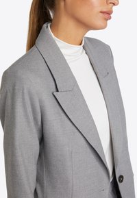 Grauer Blazer mit breiten Revers und einem einzigen Knopf, getragen über einem weißen Rollkragenpullover. Der Stoff wirkt strukturiert mit einer maßgeschneiderten Passform.