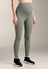 Nosečniške legice v mehki olivno zeleni barvi, visoko pasu, gladke teksture, v kombinaciji z črnimi športnimi čevlji.