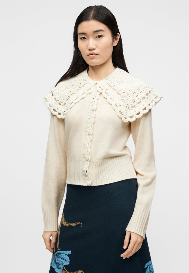 Vrouw draagt een crèmekleurig vestje met knopen, voorzien van een grote gehaakte kraag, en een donkere rok met blauwe bloemenborduursels, staand tegen een witte achtergrond.