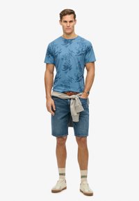 Blue short-sleeve t-shirt with dark floral print, denim shorts, grey sweater tied at waist, and light sneakers with cream and green accents.