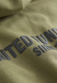 Tissu vert à la texture douce avec un texte noir caoutchouté indiquant en lettres grasses « UNITED NUNS » et « SIMPLE ».