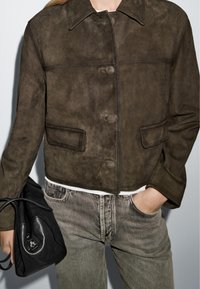 Brown suede cropped jacket with a pointed collar, front button closure, two chest pockets, and textured surface. Paired with grey denim pants.