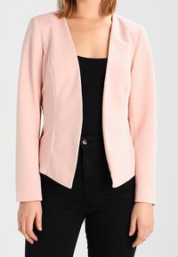 Blazer ajusté rose clair à manches longues, surface texturée, design à ouverture frontale sans fermetures visibles, associé à un haut noir.