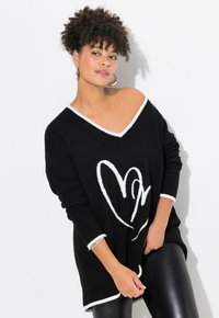 Schwarzer Pullover mit V-Ausschnitt, weißem Saum, überschnittener Passform, mit einem weißen Herzgrafik. Getragen mit schwarzen Leggings, die den Texturkontrast betonen.