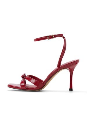 Sandalo con tacco alto in vernice rossa con dettaglio frontale annodato, due cinturini orizzontali e un cinturino alla caviglia. Punta leggermente appuntita.