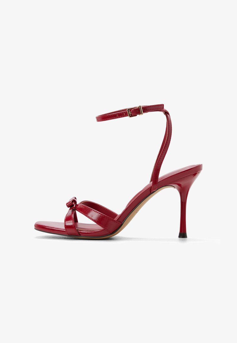 Czerwona lakierowana sandałka na wysokim obcasie z węzłowym detalem na przodzie, dwoma poziomymi paskami i pojedynczym paskiem wokół kostki. Lekko spiczasty nosek.