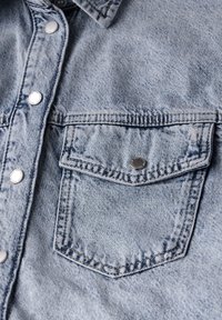 Camicia di jeans azzurro chiaro con bottone frontale, bottoni argentati e tasca sul petto. Tessuto texturizzato con dettagli di cucitura.