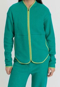 Giacca in fleece verde acqua con colletto arrotondato, zip verde e rifiniture in verde limone a contrasto. Presenta una chiusura con zip e una texture liscia.