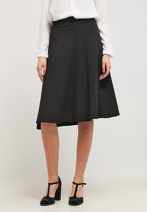 Kobieta ubrana w czarną spódnicę typu A o długości do kolan, białą bluzkę z długim rękawem oraz czarne szpilki z paskiem wokół kostki, stojąca na jednolitym tle.