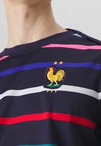 Marinblå randig t-shirt med breda horisontella ränder i röd, rosa, blå och vit. Inkluderar en broderad gul tupp och två stjärnor.