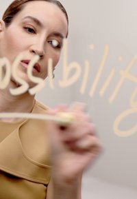 Kvinna i en beige topp som skriver "möjlighet" i guld på en slät, ljus yta med en klar penna, med ett koncentrerat uttryck.