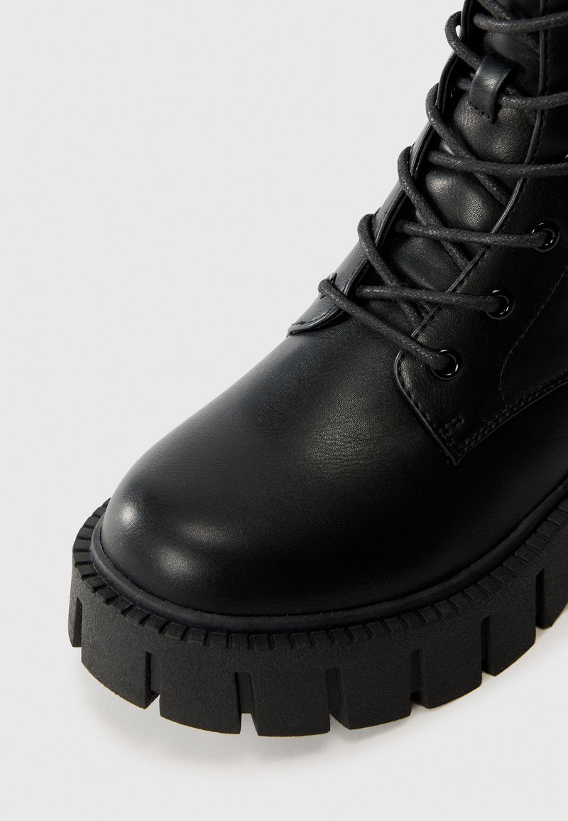 Bottine en cuir noir avec un bout rond, semelle épaisse à rainures et un laçage avec œillets en argent. Texture lisse avec des accents minimalistes.