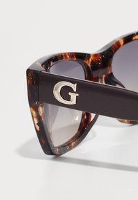  Schildpadbril met een donker montuur, vervagende glazen en een metalen "G" accent op de arm. Beschikt over een geometrisch, hoekig ontwerp.
