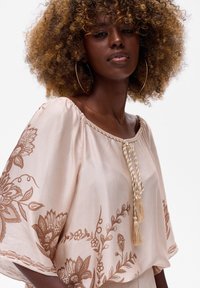 Lichtroze blouse met decoratieve bruine borduursels, brede vleermuismouwen en een geknoopte franjesdetail bij de halslijn. Gladde stoftextuur.