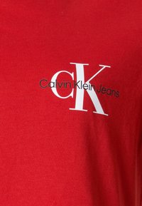 Czerwona bawełniana koszulka z brandingiem Calvin Klein Jeans. Logo zawiera "CK" w białym kolorze i "Calvin Klein Jeans" w czarnym. Miękka tekstura.