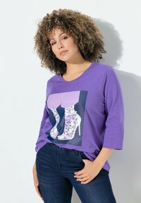 Lila t-shirt med trekvartsärmar, med en grafik av höga klackskor prydda med ett blommönster mot en mörk bakgrund.