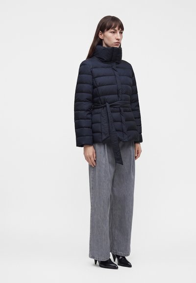 Femme portant une doudoune noire ceinturée, un pantalon large gris et des bottines noires à talons, debout sur un fond blanc.