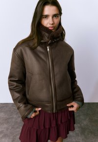 Veste en cuir marron avec un col haut doublé de fourrure, dotée d'une fermeture éclair à l'avant et de deux poches latérales, portée par-dessus une robe bordeaux à volants.