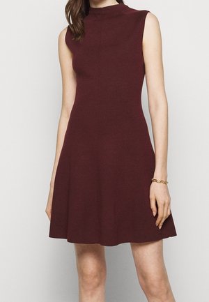 Kobieta ubrane w bezrękawnikową bordową dzianinową sukienkę z wysokim dekoltem i rozkloszowanym dołem, stojąca na tle jednolitego jasnego tła.