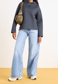 Grijze hoodie met een logo, lichtblauwe brede jeans en witte sneaker. Olijfgroene schoudertas voegt contrast toe. Gladde texturen.