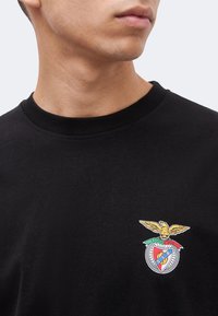 Κοντινό πλάνο ενός ατόμου που φορά μαύρο πουκάμισο με πολύχρωμο έμβλημα αετού στο στήθος, το κεφάλι γυρισμένο ελαφρώς προς το πλάι.