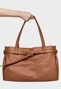 Brązowa skórzana torba typu tote z dwoma długimi uchwytami, detalem w formie paska oraz srebrnymi elementami metalowymi. Wytłoczone logo marki na dole. Gładka tekstura.