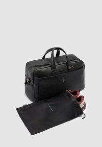 Sac de voyage en cuir noir avec poignées supérieures, présentant une surface texturée et des ferrures en bronze. Comprend une housse à chaussures noire avec un logo bleu, contenant des baskets.