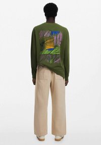 Camicia verde a maniche lunghe con un design astratto colorato sul retro. Abbinata a pantaloni beige a gamba larga e scarpe di colore chiaro.