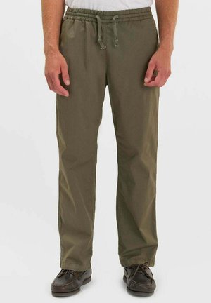 Man draagt olijfgroene broek met trekkoord en donkerbruine schoenen, staand tegen een effen witte achtergrond.