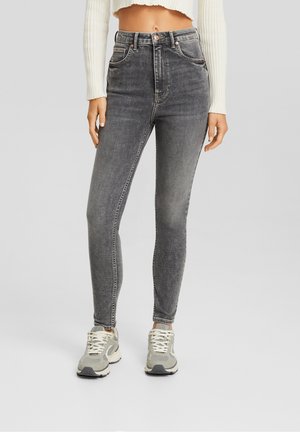Femme portant un jean skinny noir délavé taille haute avec poches avant et des baskets blanches, debout devant un fond uni.