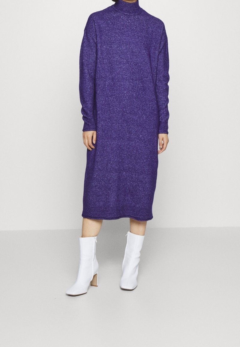 Kvinna klädd i en lila stickad turtleneck-klänning som går till knäna, med långa ärmar och vita högklackade ankelstövlar, stående på ett slätt golv.