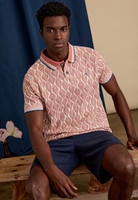 Korte mouwen polo in koraal met golfpatroon, voorzien van een lichtroze kraag en blauwe accenten, gedragen met marineblauwe shorts, zittend op een houten oppervlak.