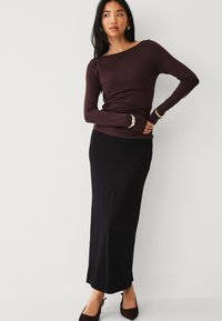 Haut à manches longues de couleur bordeaux associé à une jupe maxi noire ajustée. Tissu lisse, design près du corps et peu de détails. Chaussures à talons bruns.