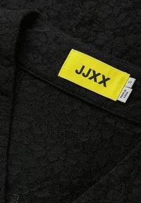 Tela de encaje negra con un patrón floral, con una etiqueta amarilla que dice "JJXX" y tamaño "S", además de información adicional de cuidado en una etiqueta blanca.