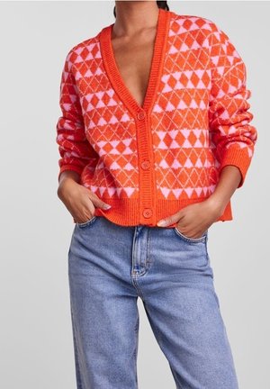 Femme portant un cardigan à motifs géométriques orange et rose avec des boutons, associé à un jean taille haute bleu clair, les mains dans les poches.