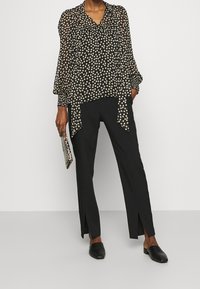 Blusa nera con pois e maniche lunghe, con scollatura a V, abbinata a pantaloni neri con fessure laterali e scarpe nere senza lacci.